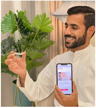 شاب سعودي يرتدي اللباس التقليدي الأبيض ويحمل هاتفه مع موقع بسمة على الشاشة وبيده الأخرى يحمل تقويم الأسنان الشفاف